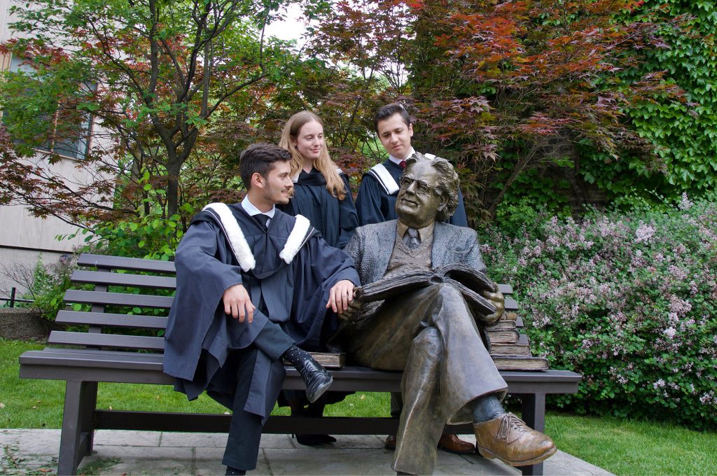 three graduates pose with sculpture of Northrop Frye