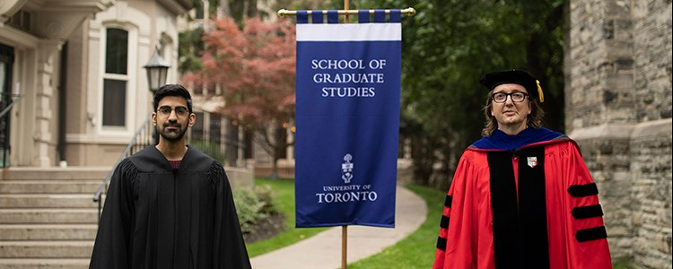 Faraz Alidina wears a black robe and stands to the left of a sign saying School of Graduate Studies. To the right of the sign is Prof. Joshua Barker, who is in red-and-black regalia for convocation.