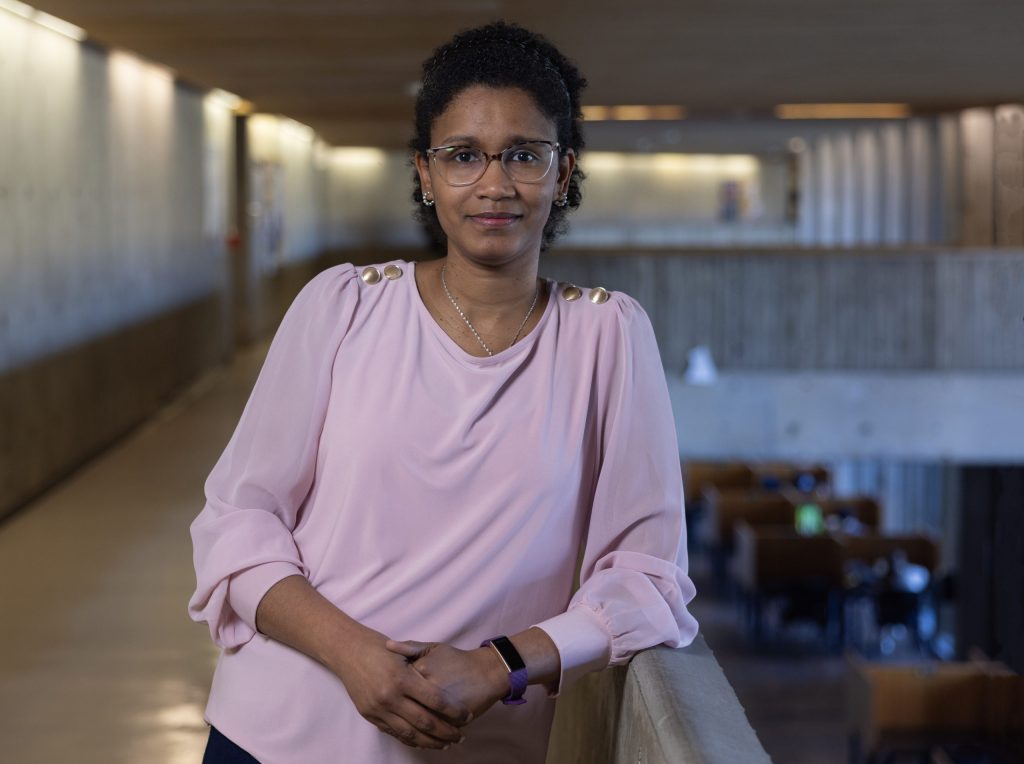 A researcher is learning against a railing, wearing a pink, long-sleeve shirt and glasses.