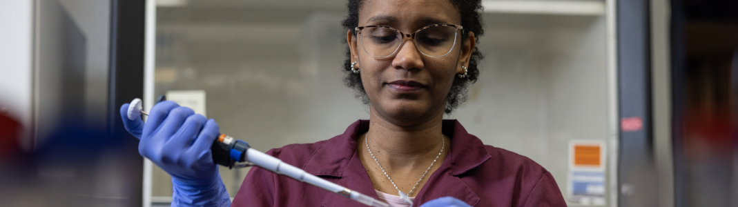 A Black woman is in a lab, wearing a purple lab coat and rubber gloves. She is wearing glasses and has short, black hair.