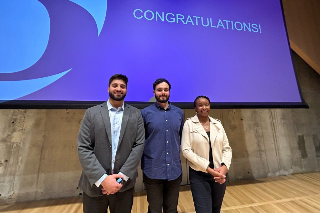 Three PhD students stand together on a stage. Behind them is a blue screen with the word CONGRATULATIONS on it.