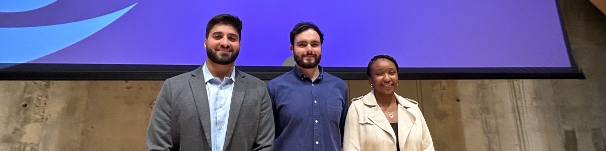 Three PhD students stand together on a stage. Behind them is a blue screen with the word CONGRATULATIONS on it.