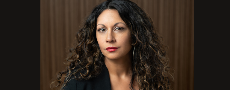 Headshot of Dr. Sharma. She has long brown hair and is wearing a dark jacket. She is pictured in front of a dark brown background.