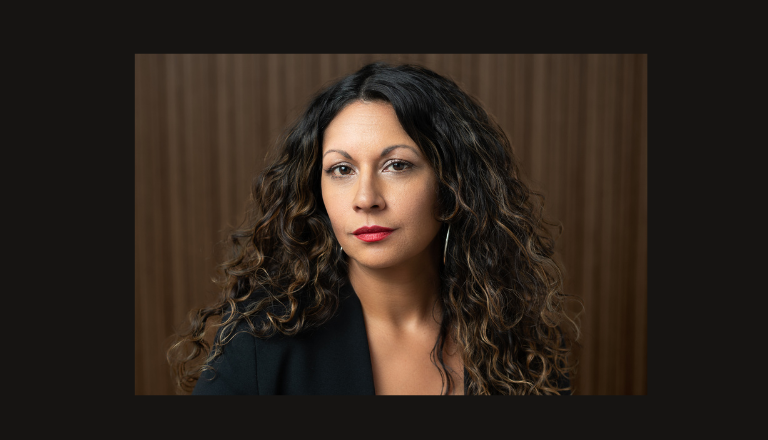Headshot of Dr. Sharma. She has long brown hair and is wearing a dark jacket. She is pictured in front of a dark brown background.