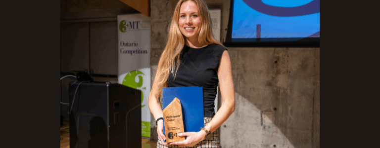 Emily Wood is standing on the 3MT stage holding her trophy. She is wearing a black top and smiling.